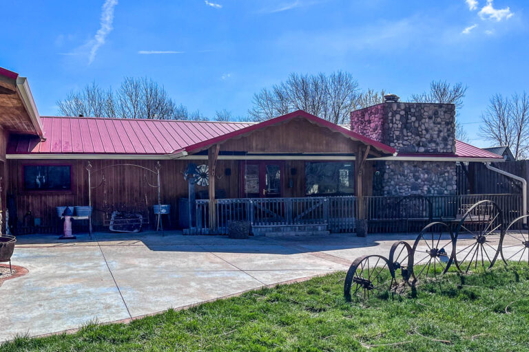 Front porch of rustic building with red roof and new Almond gutters installed, wagon wheels displayed in the yard.