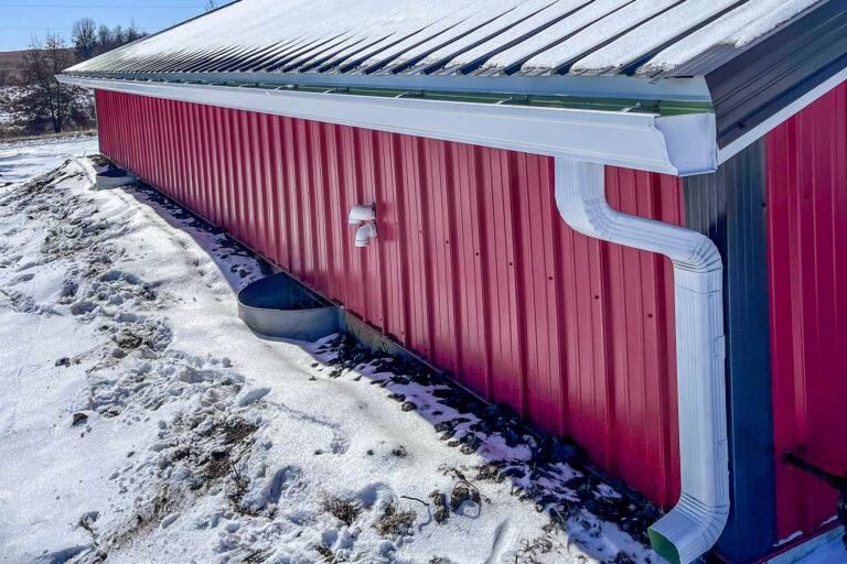 Side view of a large red metal building with a snow-covered roof and new white 6-inch gutters installed.