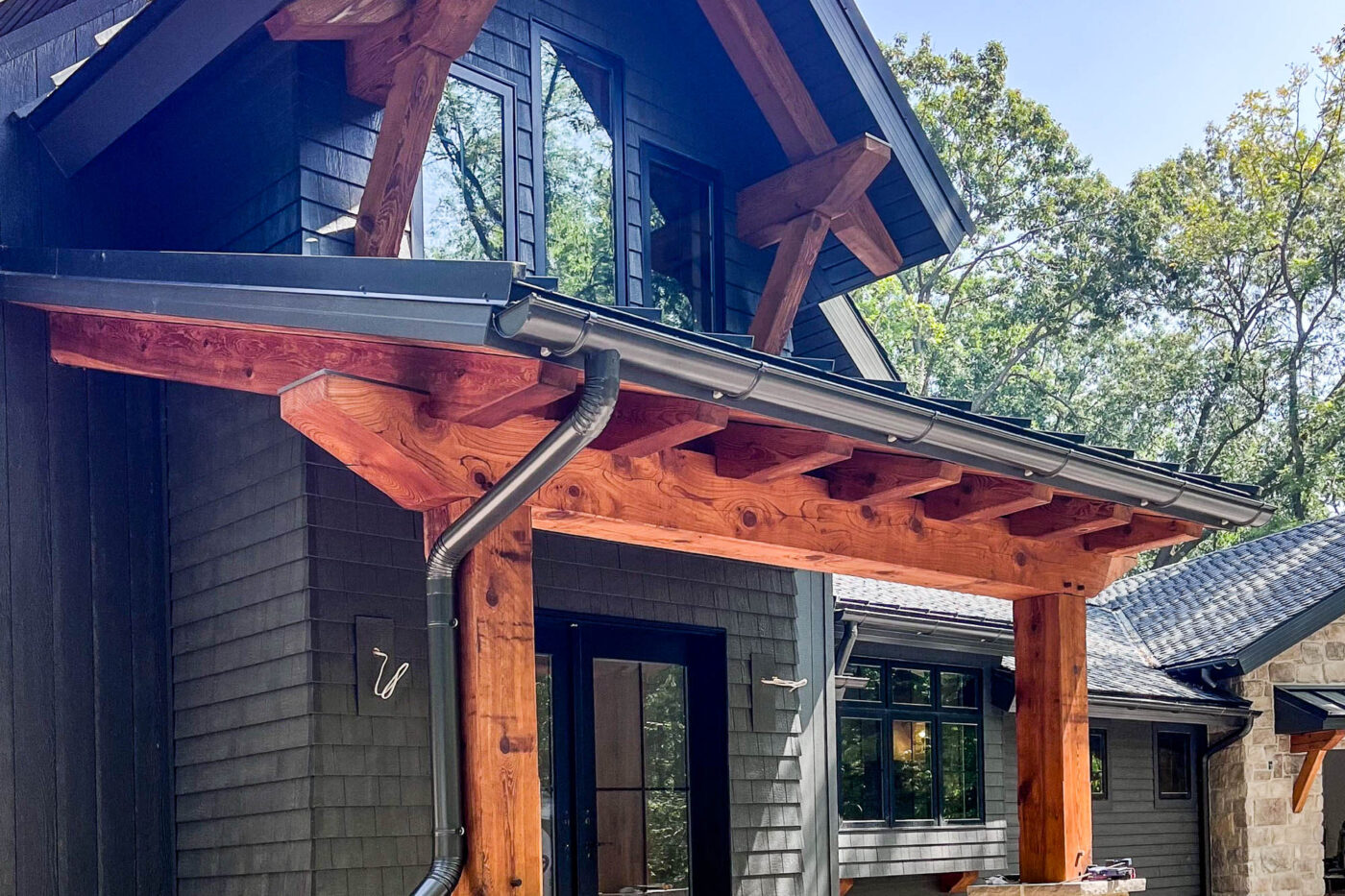 Round bronze downspout installation on timber and stone entryway in Kearney, MO, combining durability and visual harmony.