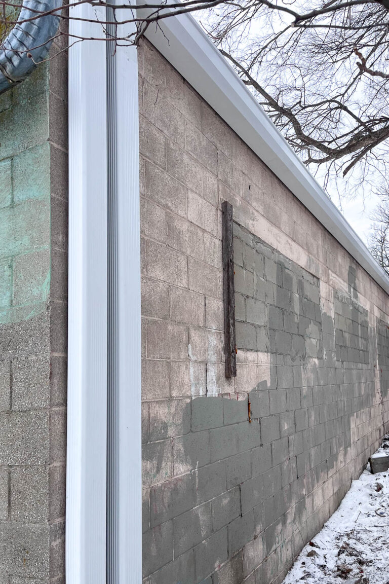 Close-up side view of a 7-inch white commercial-grade gutter and large 4x5 downspout securely mounted on a weathered concrete block wall.