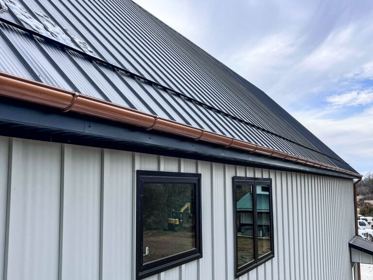Close-up view of a standing seam metal roof with 6-inch half-round copper gutters and a round copper downspout mounted on a light-colored modern exterior wall.