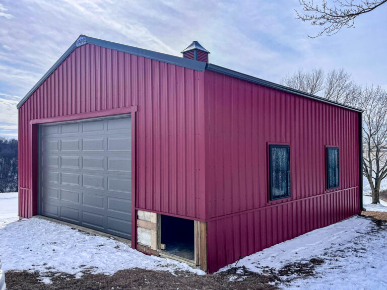 A red metal building with a snow-covered ground, featuring a recently installed 5-inch Musket Brown aluminum gutter system with matching 3x4 downspouts on the side. The building has a large gray garage door, black-framed windows, and a visible downspout running vertically along the corner.