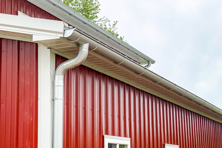 Angled view of galvanized half-round gutters and downspouts installed on a red metal building in Rosebud, Missouri.