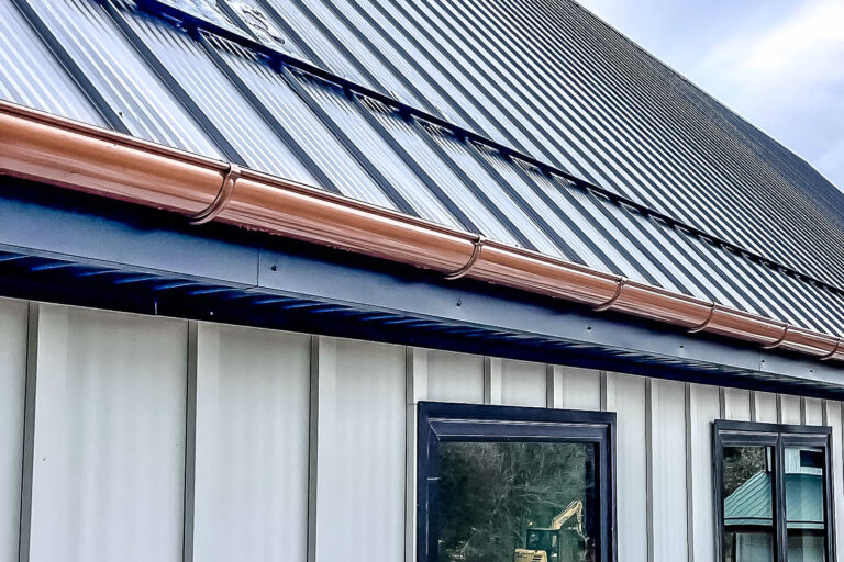 Copper gutters installed on a white metal building with a standing seam metal roof, reflecting natural light beneath a partly cloudy sky.