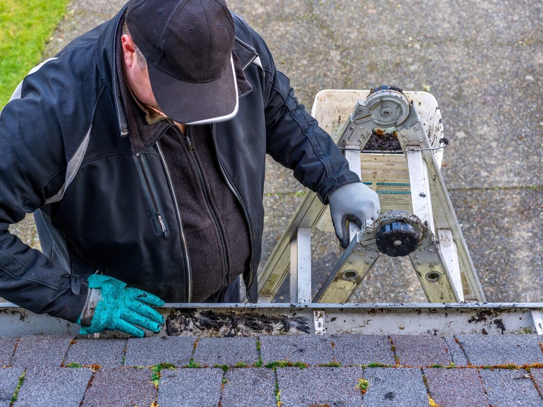 man on top of ladder cleaning dirt from roof gutter on building his hand in messy gloves view from the top of the roof